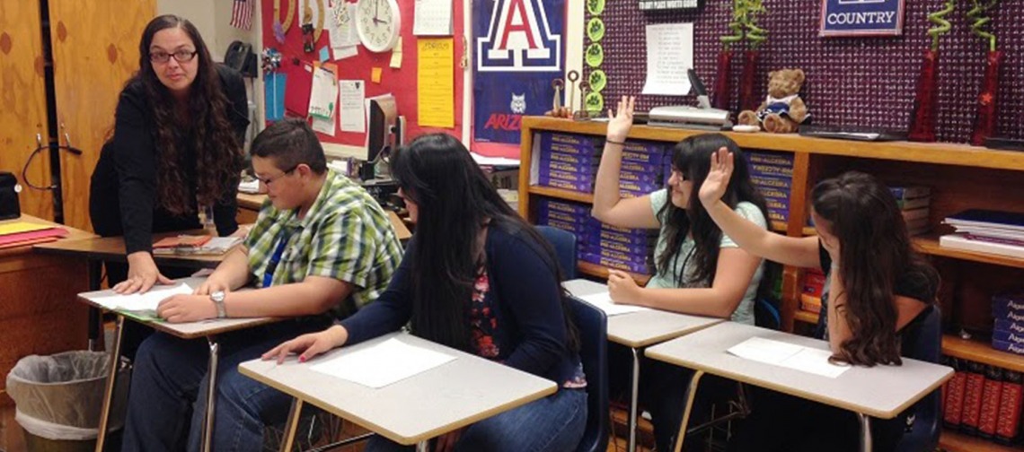 four students sitting at their desk, teacher in the background, two students raising their hands