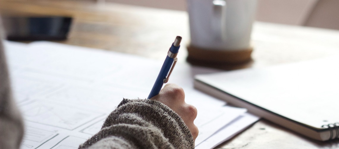 hand holding a pen writing in a notebook, coffee mug in the background